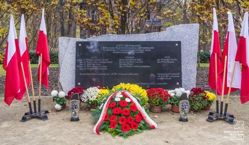 Pomnik kamienny z tablicą marmurową, po prawej i lewej stronie flagi Polskie na drzewcach w stojakach, przed pomnikiem kwiaty i znicze
