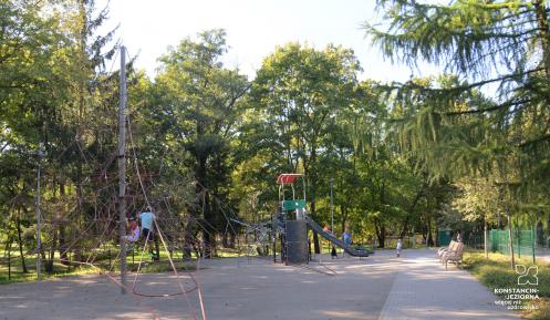  Plac zabaw z dwoma dużymi urządzeniami: do wspinania oraz zjeżdżania. Bawi się na nim kilkoro dzieci. Wokół rosną wysokie drzewa. 