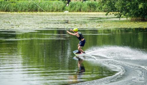 Staw, na środku którego mężczyzna uprawia wakeboarding. Ma na sobie kamizelkę ratunkową, kask na głowie, na nogach przypiętą podłużną deskę, dzięki której może ślizgać się po tafli wody. W rękach trzyma uchwyt połączony z liną, która go ciągnie. 