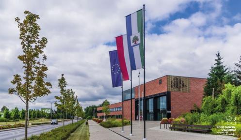 Duży, nowoczesny budynek użyteczności publicznej  ukazany z dużej perspektywy narożnika, ceglane ściany, duże okna i oszklenia, przed budynkiem drzewa oraz horągiew Konstancina-Jeziorny, flagi Polski i Unii Europejskiej na masztach 