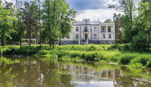 Dwupiętrowy, symetryczny budynek na planie prostokąta z jasnoszarą elewacją w otulinie parku i rzeki.