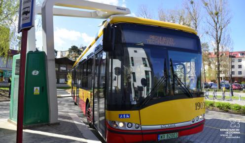Żółto-czerwony autobus stoi pod wysięgnikiem stacji do ładowania. Z lewej strony znak drogowy. Tło stanowi zabudowa wielorodzinna.