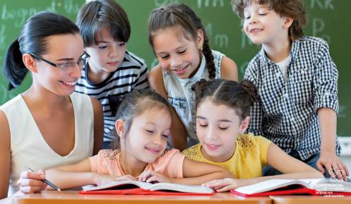 Pięcioro uczniów ogląda z nauczycielką dwie  otwarte książki położone na biurku, w tle zielona tablica szkolna.