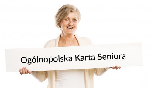 Na zdjęciu uśmiechnięta seniorka, która w rękach trzyma dużą kartę papieru, na której znajduje się napis - Ogólnopolska Karta Seniora. 