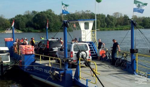 Fragment rzeki Wisły z promem, na którym stoją dwa samochody oraz kilka osób. W powietrzu  powiewają flagi gminy Konstancin-Jeziorna