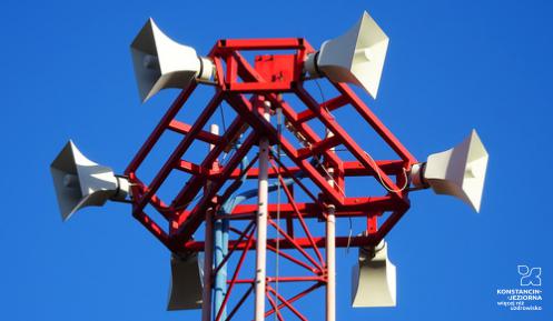 sześć białych głośników syreny alarmowej ułożonych po okręgu na czerwonej konstrukcji, wzniesione wysoko na tle niebieskiego nieba.