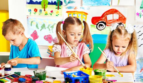 Dwie dziewczynki i chłopczyk siedzą przy stole i malują farbami prace plastyczne. Przed nimi słoiczki z kolorowymi farbami. W tle na tablicy wiszą kolorowe rysunki.