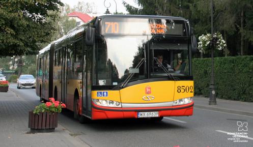 Przód autobusu miejskiego linii 710, jadącego po ulicy, z prawej strony chodnik na którym stoją donice z kwiatami i latarnie