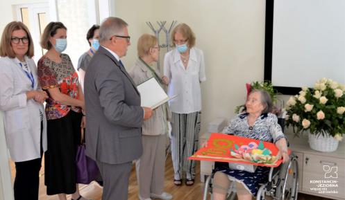 W głównej części mężczyzna, który stoi i trzyma w ręku list gratulacyjny. Obok niego w sali, na wózku inwalidzkim, siedzi starsza kobieta. W tle widać kosze kwiatów oraz inne stojące osoby. 