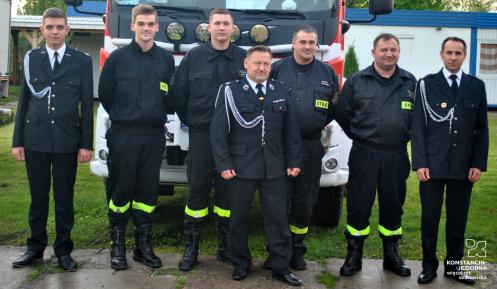 Siedmiu mężczyzn stoi w mundurach strażackich na tle wozu strażackiego.