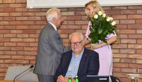 Na pierwszym planie mężczyzna w czarnej marynarce siedzi przy stole, za nim kobieta w różowej sukience (przewodnicząca rady) wręcza kwiaty mężczyźnie w szarym garniturze – burmistrzowi gminy.