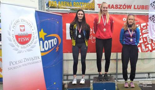 Podium, na nim stoją 3 młode dziewczyny. Na podeście z numerem 1 stoi – zawodniczka  Uczniowskiego Klubu Sportowego Zryw – Wiktoria Dąbrowska. W tle widać banery reklamowe. 