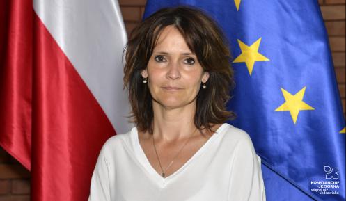 Zdjęcie portretowe kobiety – ciemne proste włosy, ubrana w jasną bluzkę. W tle flaga Polski i Unii Europejskiej.