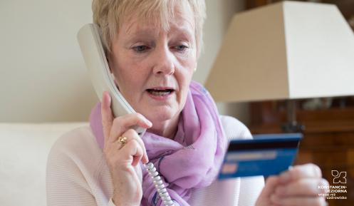 Starsza kobieta rozmawia przez telefon stacjonarny, w jednym ręku trzyma słuchawkę telefonu stacjonarnego, w drugim – kartę bankomatową.