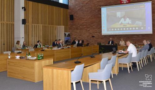 Sala konferencyjna ratusza. Przy trzech prostokątnych stołach, ustawionych w odwróconą literę U, stoi po dłuższych bokach kilka beżowych krzeseł, na niektórych siedzi kilkanaście osób. Na ścianie z czerwonej cegły wisi ekran, na którym wyświetlany jest obraz z sali.