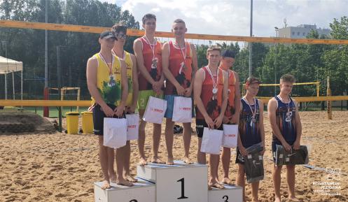 Podium sportowe. Na podestach z numerem 1,2,3 stoją w parach zawodnicy. Na szyi mają medale. W rękach torby z upominkami. W tle widać boisko do siatkówki plażowej. 