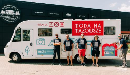 Pięciu mężczyzn, w czarno-białych koszulkach i okularach przeciwsłonecznych, stoi na tle samochodu typu kamper z logo Moda na Mazowsze. Jeden z nich wskazuje na napis: #KamperemPoMazowszu.