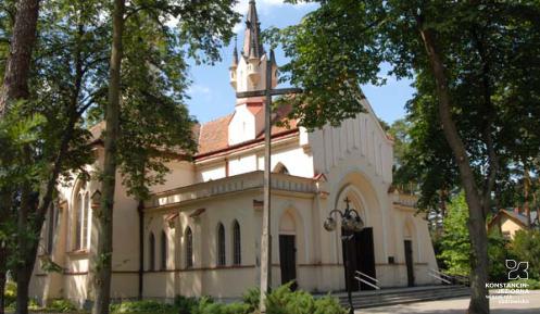 Budynek kościoła parafialnego z portalem, nawą główną, wieżą i czerwoną dachówką w otoczeniu drzew.