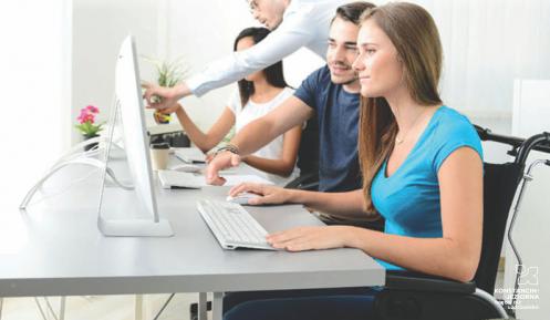 Cztery osoby w tym dwie kobiety, z których jedna jest na wózku inwalidzkim, patrzą w monitory komputerowe. Mężczyźni tłumaczą coś kobietom, jeden z nich palcem dotyka ekranu monitora. Na biurku leżą klawiatury i myszki komputerowe, stoi kwiat i leżą dokumenty.