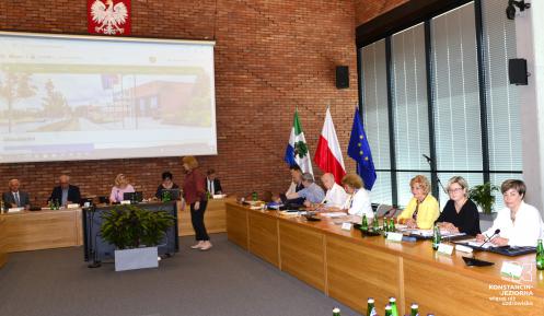 Sala konferencyjna ratusza. Przy dwóch stołach ustawionych – jeden pod oknem, drugi pod ścianą z czerwonej cegły – siedzi grupa osób (kobiet i mężczyzn). Na ścianie wisi ekran, na którym wyświetlana jest strona internetowa gminy Konstancin-Jeziorna. 