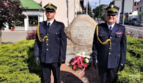 Dwóch strażników miejskich, ubranych w stroje galowe, stoi na baczność obok pomnika upamiętniającego bohaterów Bitwy Warszawskiej z 1920 roku. Pod pomnikiem zostały złożone biało czerwone kwiaty.