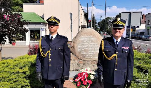 Dwóch strażników miejskich, ubranych w stroje galowe, stoi na baczność obok pomnika upamiętniającego bohaterów Bitwy Warszawskiej z 1920 roku. Pod pomnikiem zostały złożone biało czerwone kwiaty.