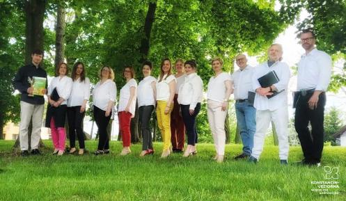 Grupa trzynastu osób. Dziewięć kobiet i czterech mężczyzn. Stoją oni obok siebie. W tle widać drzewa.
