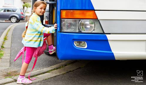 Dziewczynka ubrana w różową sukienkę i zielony sweter wsiada do biało-niebieskiego autobusu.