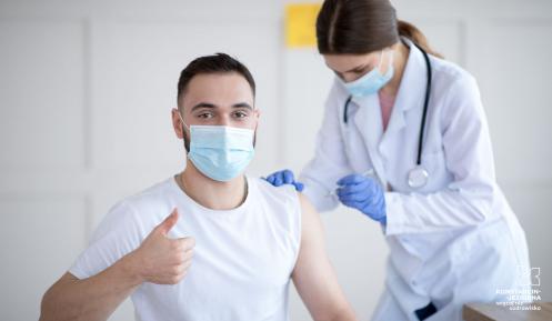 W gabinecie lekarskim młody mężczyzna w maseczce ochronnej otrzymuje zastrzyk, który wykonuje lekarka ubrany w biały fartuch, na szyi ma stetoskop.