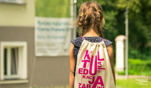 Kilkuletnia dziewczynka idzie po trawie. Na plecach ma jasny worek z napisem kultura, edukacja i zabawa. 