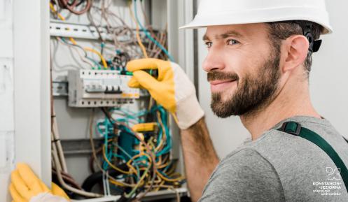 Pomieszczenie gospodarcze, na ścianie zamontowana jest skrzynka elektryczna. Po prawej stronie stoi mężczyzna - elektryk, który sprawdza bezpieczniki. 