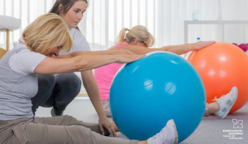 Sala fitness. Dwie kobiety siedzą na podłodze, przed sobą mają duże kolorowe piłki. Wykonują ćwiczenia. Między nimi stoi instruktorka. 