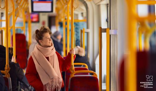 Dorosła kobieta ubrana w jasną kurtkę i szalik na szyi stoi w autobusie. Trzyma się żółtej barierki. 