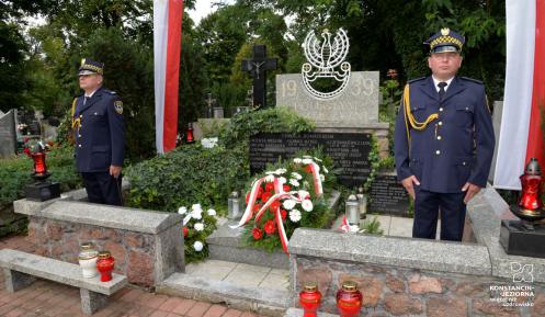 Cmentarz. Przy pomniku ofiar drugiej wojny światowej stoi dwóch mężczyzn w średnim wieku. Ubrani są w mundury Straży Miejskiej. Na pomniku leżą cztery wieńce z biało-czerwonymi kwiatami. Przed pomnikiem stoją cztery znicze, trzy czerwone i jeden biały. Po obu stronach pomnika na sztandarze wiszą biało-czerwone flagi. 