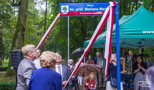 Trzy osoby dokonujące odsłonięcia umieszczonej na pionowym slupie poziomej tabliczki z nazwą „Skwer im. prof. Mariana Weissa”, kobieta w niebieskiej sukience widoczna tyłem, po jej lewej stronie młody mężczyzna w garniturze, na wprost starszy mężczyzna w garniturze, wszyscy pociągają za końcówki biało-czerwonej szarfy, obok stoi dziecko pod parasolem, w tle duży namiot z zielonym dachem, pod którym stoją inne osoby.