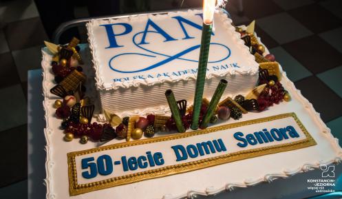 Na stoliku stoi duży biały tort z napisem Polska Akademia Nauk oraz 50-lecie Domu Seniora. Dookoła są czekoladowe ozdoby.