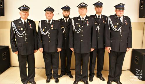 6 mężczyzn ubranych w galowe mundury strażackie stoi obok siebie w rzędzie. W tle widać kremową ścianę budynku. 