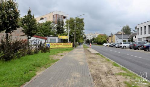 Miasto. Po lewej stronie rośną: zielona trawa, krzewy i drzewa, za nimi są budynki. Obok znajduje się chodnik, po prawej stronie jest ulica dwupasmowa, wzdłuż niej stoją zaparkowane samochody. W oddali są budynki. W tle pochmurne niebo.