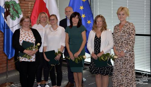  Sala konferencyjna. Siedem osób stoi obok siebie w rzędzie – sześć kobiet i jeden mężczyzna. Cztery kobiety w rękach trzymają dyplomy i białe róże. W tle trzy flagi: Konstancina-Jeziorny, Polski i Unii Europejskiej.