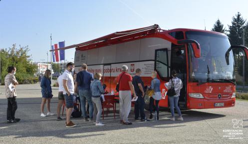 Miasto. Na chodniku stoi czerwony autokar, z boku jest duży napis Mobilny punkt poboru krwi, obok jest godło Polski i napis Minister Zdrowia. Przed autokarem w kolejce stoi dziesięć osób.