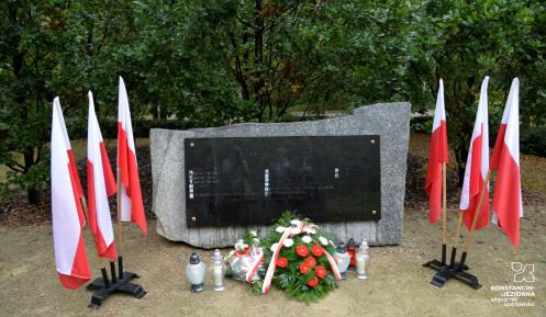 Park. Pomnik ofiar katyńskich. Przy nim stoją znicze oraz leżą: biało-czerwone kwiaty i wiązanka z białymi kwiatami. Po obu stronach pomnika stoją na stojaku trzy biało-czerwone flagi. W tle zielone drzewa.