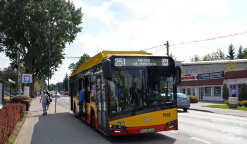 Ulica. Przy przystanku obok chodnika stoi żółto-czerwony autobus komunikacji miejskiej. Z przodu na górze jest napis 251 Metro Wilanowska. Do autobusu wsiada mężczyzna. Chodnikiem idą dwie kobiety.