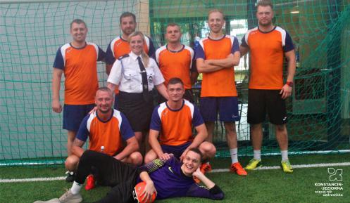 Zdjęcie grupowe drużyny piłkarskiej. 8 zawodników w pomarańczowych koszulkach stoi w bramce. Między nimi jest jedna kobieta ubrana w mundur galowy Ochotniczej Straży Pożarnej. 