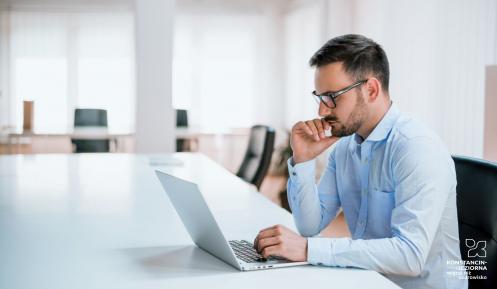 Biuro. Młody mężczyzna siedzi na krześle przy białym biurku. Ubrany jest w błękitną koszulę, na nosie ma okulary. Przed nim stoi srebrny laptop.