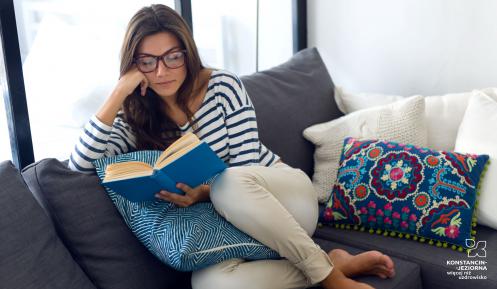 Młoda kobieta siedzi na kanapie, w ręku trzyma i czyta książkę, na nosie ma okulary. Na kanapie leżą poduszki.