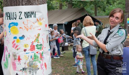 Kobieta w mundurze harcerskim gestem rąk zaprasza do obozu harcerskiego, w tle kilkadziesiąt osób oraz zielony namiot. Na drzewie wisi kolorowy obrazek z napisem: Maluj z nami.