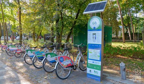 Stacja wypożyczalni rowerów miejskich z sześcioma rowerami. W tle drzewa, ulica z przejeżdżającymi samochodami oraz lokomobila.   