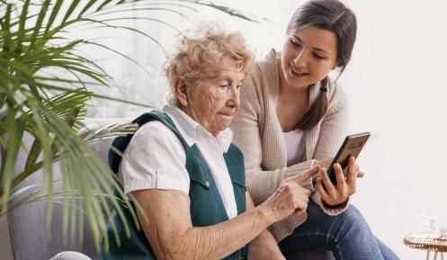 Pokój. Na fotelu siedzi seniorka, obok siedzi młoda dziewczyna. Dziewczyna w ręku trzyma telefon. Seniorka pokazuje palcem coś w telefonie.