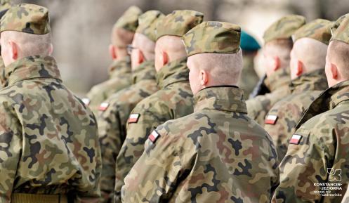 Ośmiu mężczyzn stoi obok siebie. Wszyscy ubrani są w wojskowe mundury, na głowach mają czapki, przy ramieniu naszyta jest flaga Polski. Widoczne są tylko ich plecy.