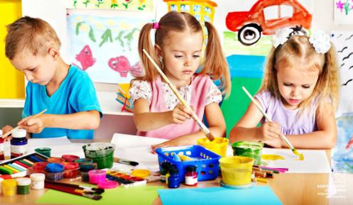 Sala przedszkolna, przy stoliku siedzą dzieci: dwie dziewczynki i jeden chłopiec. Na stole leżą farby do malowania. Wszyscy trzymają pędzle w rękach i coś tworzą. 